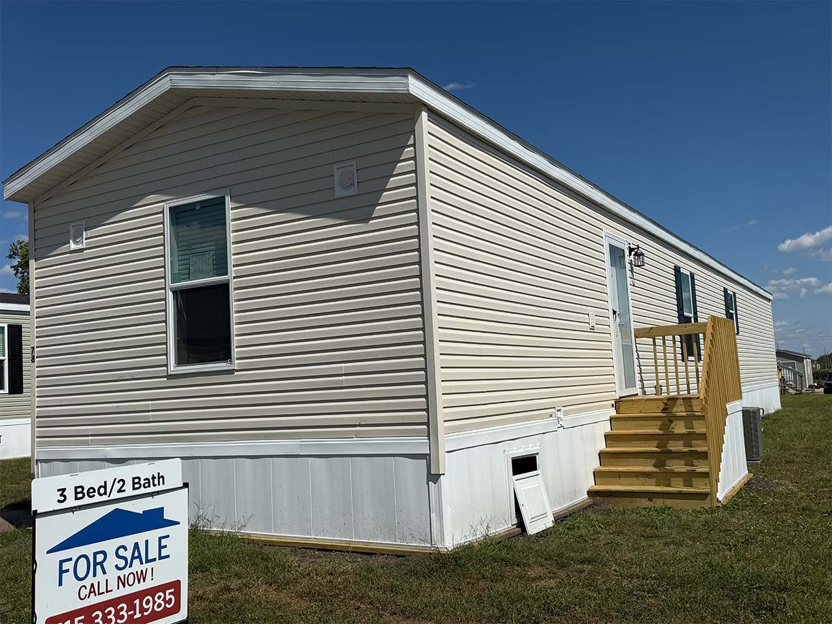 A beige manufactured home with white trim and a wooden staircase at the entrance is shown on a grassy lot. A "For Sale" sign in front advertises 3 bedrooms, 2 baths, and displays a phone number.