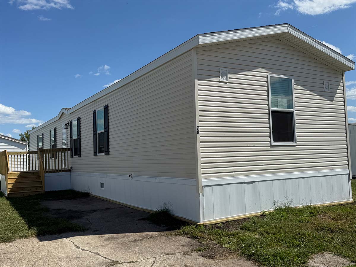 A beige mobile home with black shuttered windows and a wooden porch, situated next to a paved driveway and grassy lawn under a blue sky with scattered clouds.