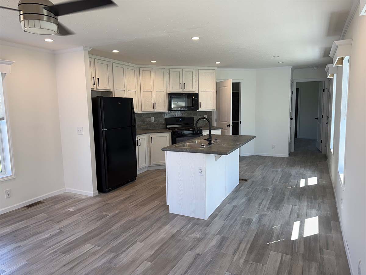A modern kitchen with white cabinets, black appliances, a center island, gray wood-look flooring, and recessed lighting. Sunlight streams through windows, brightening the open layout. A hallway leads to other rooms.