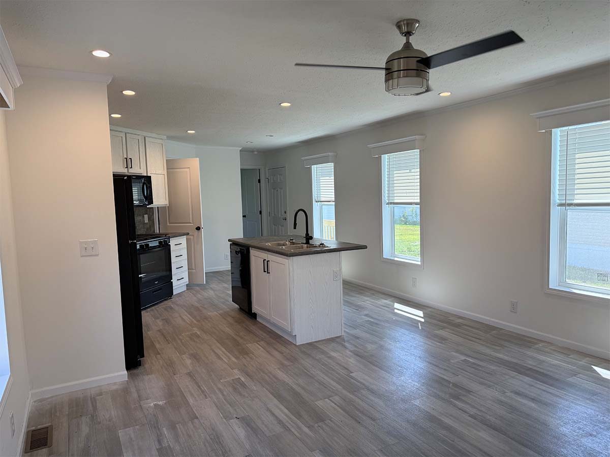 Modern, unfurnished kitchen and living area with light wood flooring, white walls, an island in the center, black appliances, ceiling fan, and multiple windows letting in natural light.