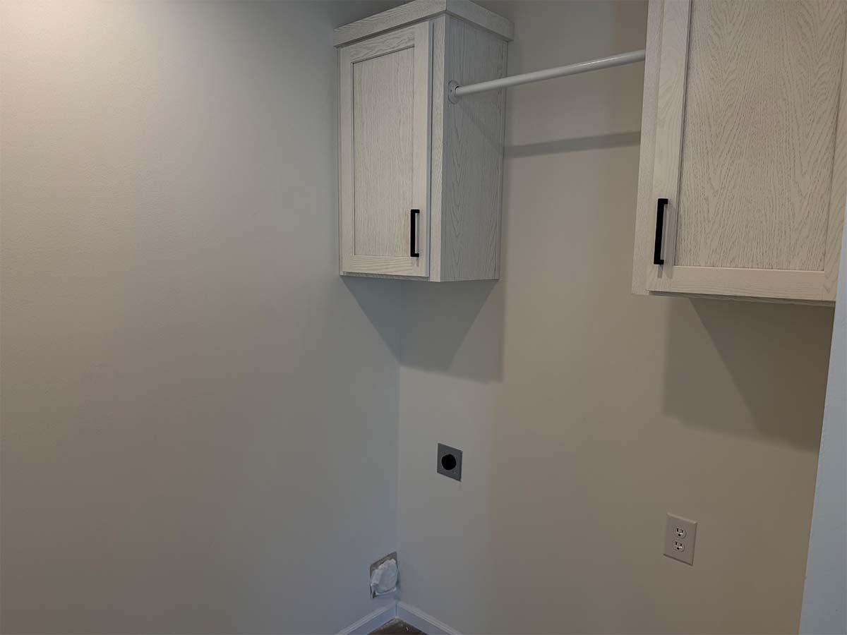 A small laundry room with light-colored walls, two light wood cabinets with black handles, a horizontal rod for hanging clothes, and connections for a washer and dryer. The floor is bare.