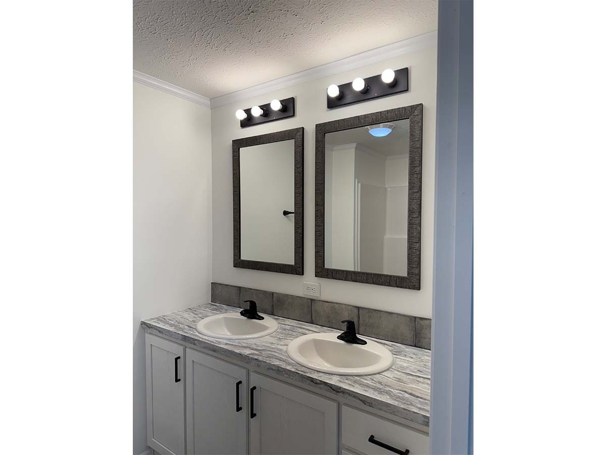 A bathroom vanity with two sinks, two rectangular mirrors with dark frames, and two sets of wall-mounted lights above the mirrors. The countertop is gray and white, and cabinets are white with black handles.