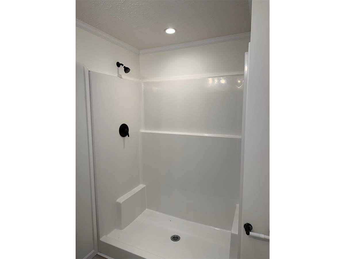 A white, walk-in shower with a built-in bench, wall-mounted black showerhead, and simple, clean walls. The shower area is well-lit by a ceiling light above.