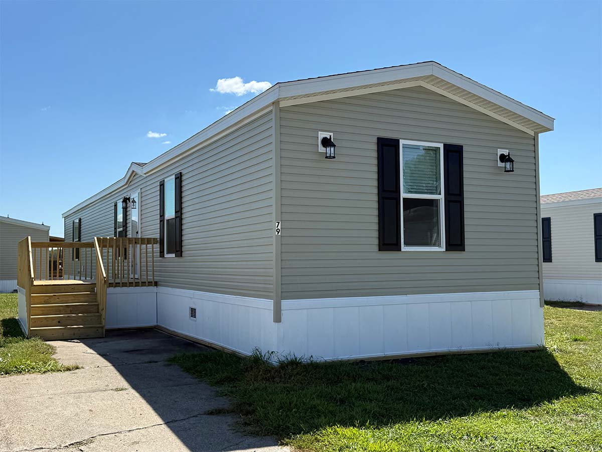A beige manufactured home with black shutters and two exterior lights. There is a small wooden porch with steps leading to the entrance, and the home sits on a grassy lot with a concrete walkway.