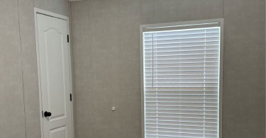 A closed white door and a window with closed white blinds are set in a beige wall. The room appears empty and has simple, neutral decor.