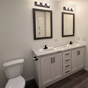 Bathroom with a double-sink vanity, two framed mirrors, overhead lighting, white cabinets, and a toilet. The walls are white and the floor is dark wood.